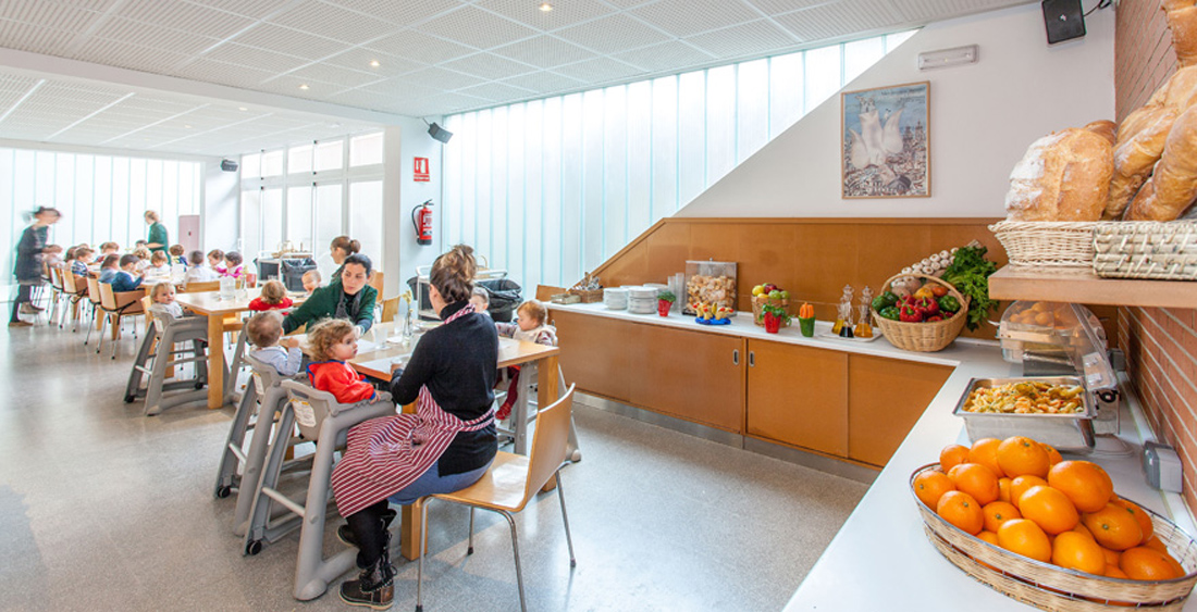 Comedor escolar del Centro Educativo Gençana. En primer plano, el buffet con fruta, hortaliza, pan y pasta. En segundo plano, los alumnos de primer ciclo de Educación Infantil comen con la ayuda de sus maestras.