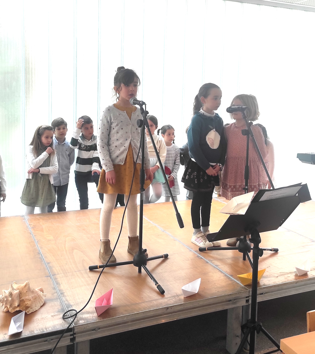 Celebración del Día de la Poesía en el Centro Educativo Gençana. Tres niñas, alumnas de 2º Educación Primaria, subidas a un escenario recitan poesía delante de dos micros. Detrás, otros compañeros las observan