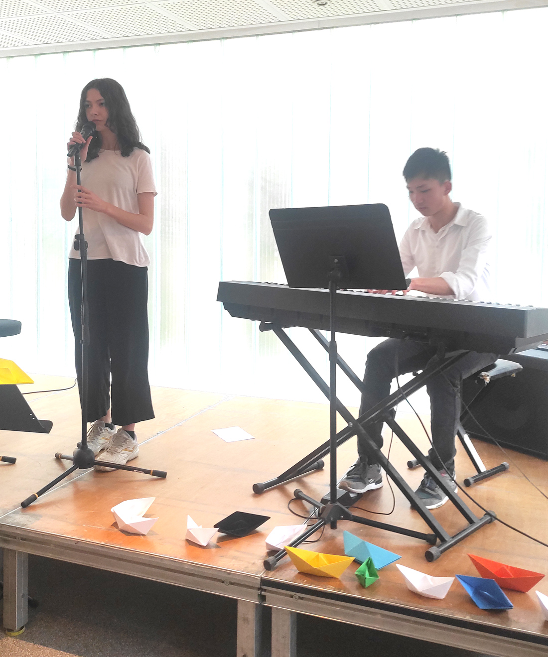 Alumnos de 3º Educación Secundaria durante la celebración del Día de la Poesía en el Centro Educativo Gençana. Una joven recita poesia de pie ante un micro y un joven toca el teclado sentado. 