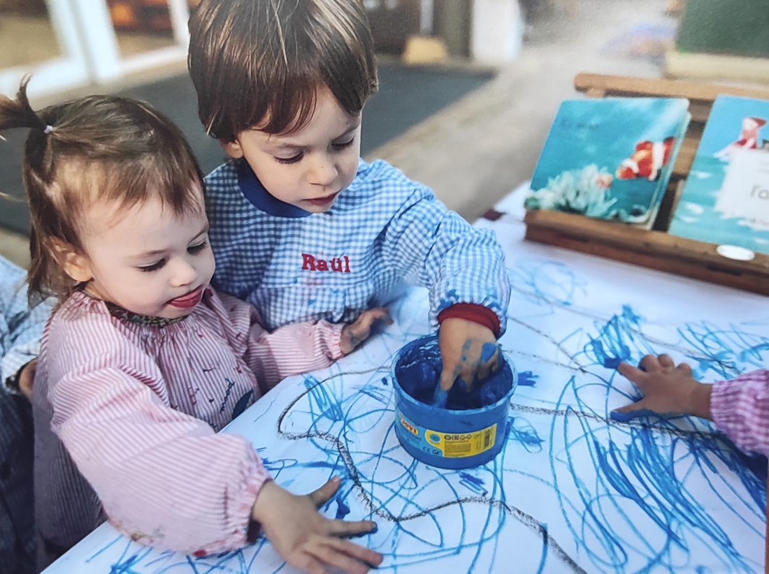 Dos niños pequeños ataviados con baby pintan con los dedos sobre una hoja blanca, mientras uno de ellos saca la lengua concentrado en su tarea