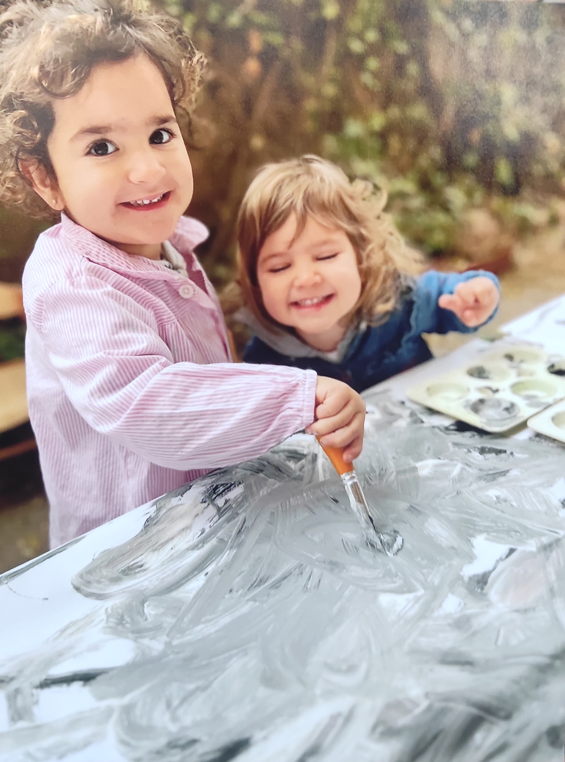 Dos niñas de la clase de 2 años del Centro Educativo Gençana pintan con pintura gris sobre una hoja en blanco mientras sonríen a cámara