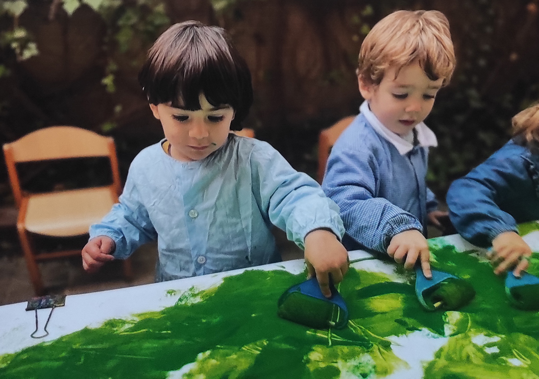 Dos niños de la clase de 2 años del Centro Educativo Gençana pintan con pintura verde utilizando rodillo sobre una hoja de papel continuo blanco