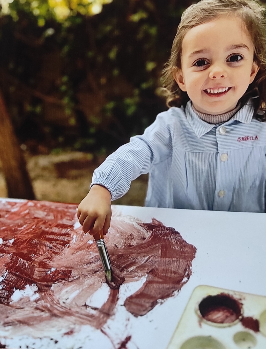 Niña pintando con tempera en color marrón sobre una hoja blanca, sonríe a cámara