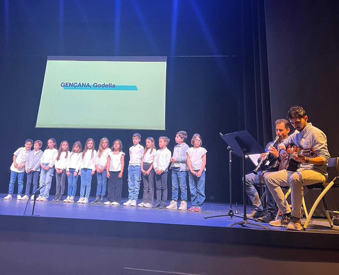 Un grupo de alumnos y alumnas de 3º Primaria del Centro Educativo Gençana cantan una canción en el escenario, acompañados al instrumento por sus profesores de Música, durante la celebración de la XII Trobada Musical de l'Horta Nord