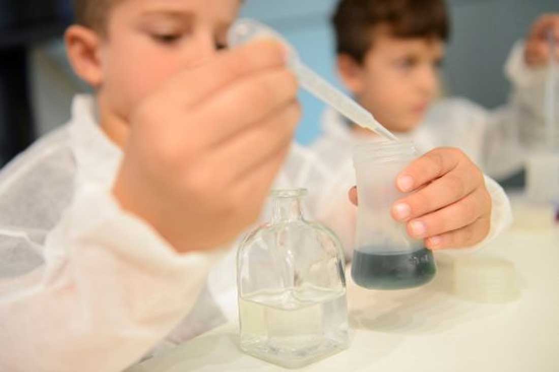 Dos niños vestidos con bata blanca introducen un líquido azul en una probeta, en un laboratorio