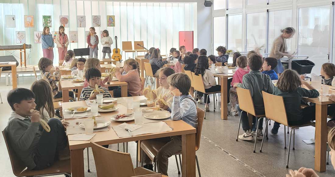 Alumnos de 2º Primaria del Centro Educativo Gençana recitan poesía subidos a un escenario en el comedor de la escuela mientras sus compañeros comen